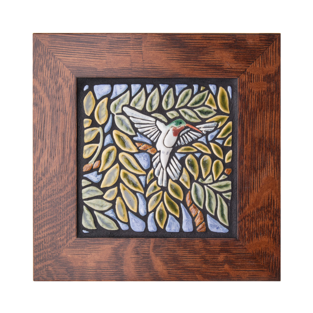 Expertly handcrafted in an artisan workshop in Canton, Ohio, this delightful ceramic tile features a colorful relief of a hummingbird in flight, surrounded by verdant leaf branches. The tile is surrounded by a high quality, 1.5" quarter sawn oak frame.  Size including frame: 10" x 10".