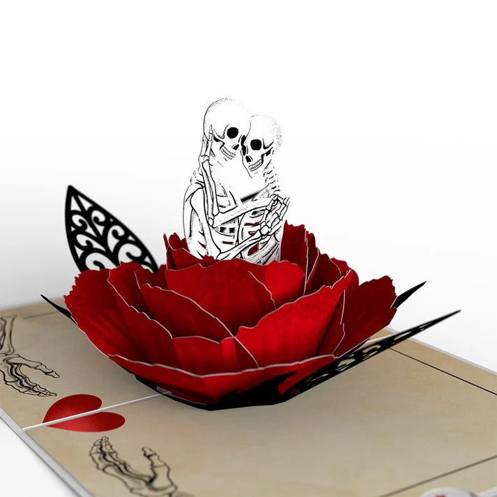 A 3D pop-up greeting card with a red background showing a skeleton couple and the text 'I am DEAD in love with you'. The card is partially opened to reveal the pop-up design.