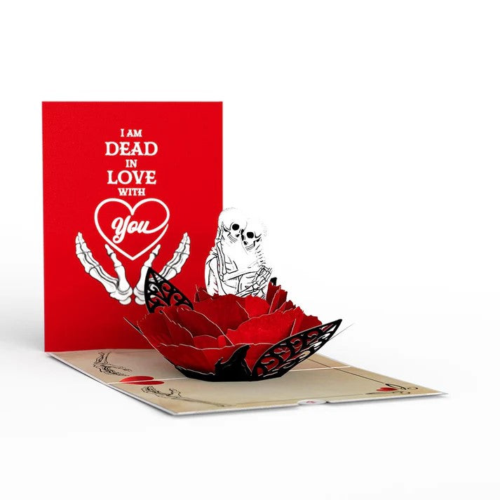 A 3D pop-up greeting card with a red background showing a skeleton couple and the text 'I am DEAD in love with you'. The card is partially opened to reveal the pop-up design.