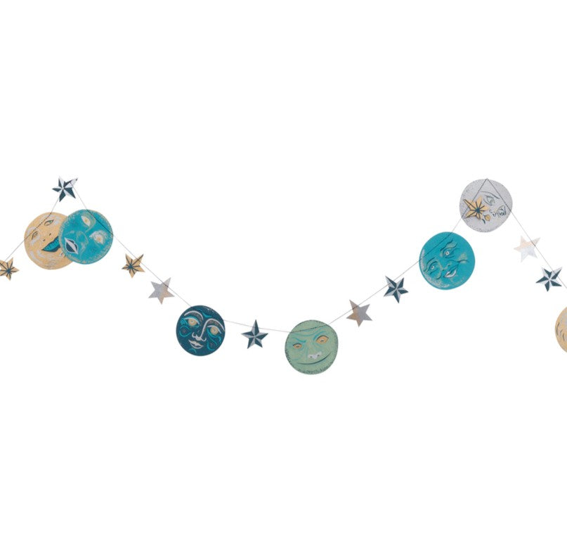A celestial-themed garland banner featuring painted moon and star shapes in teal, blue, beige, and silver, strung along a thin white cord against a plain white background.