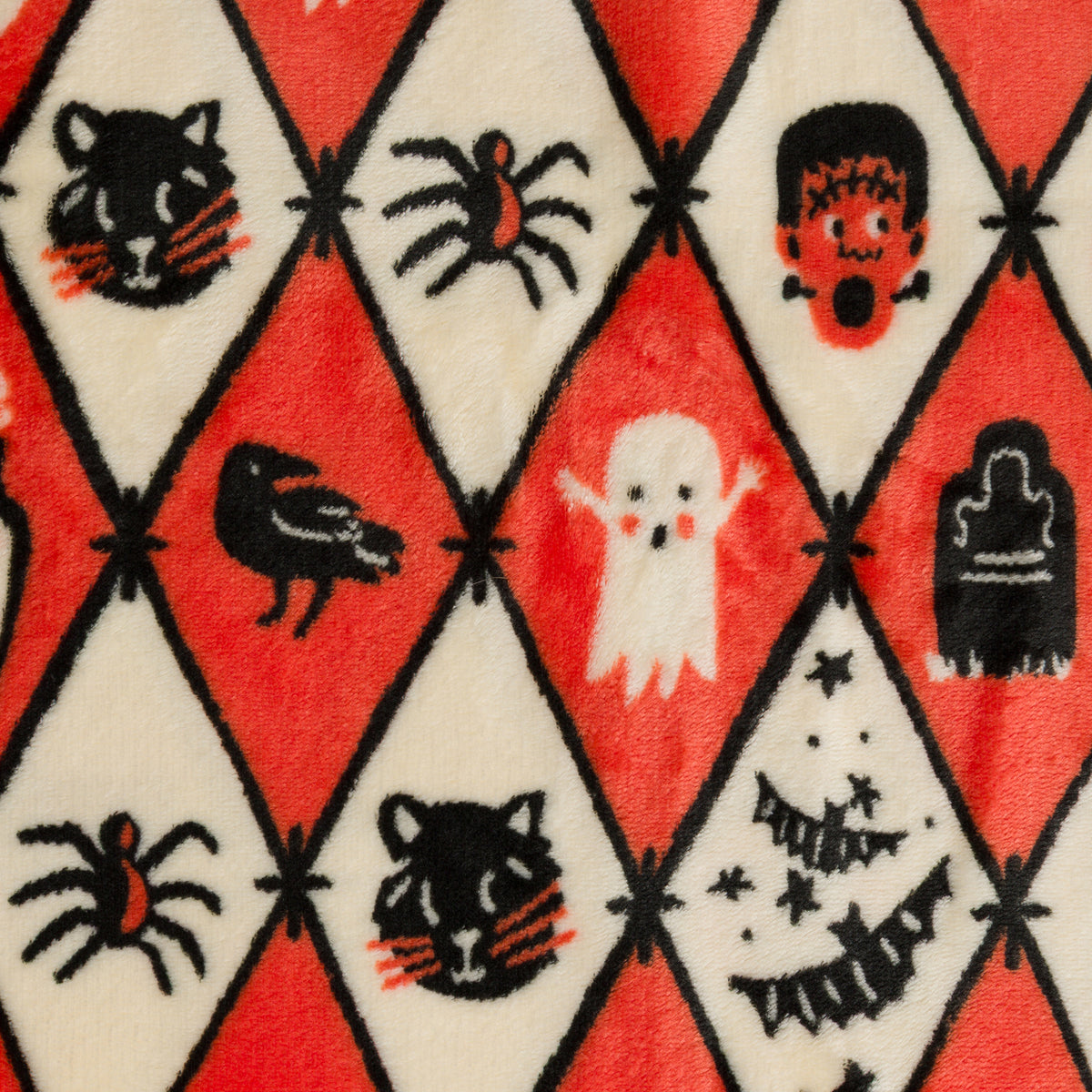 A close-up of a plush fabric with a Halloween harlequin pattern featuring red, cream, black, and orange motifs including spiderwebs, a cat face, a spider, a bird, a ghost, a tombstone, and a Christmas tree.