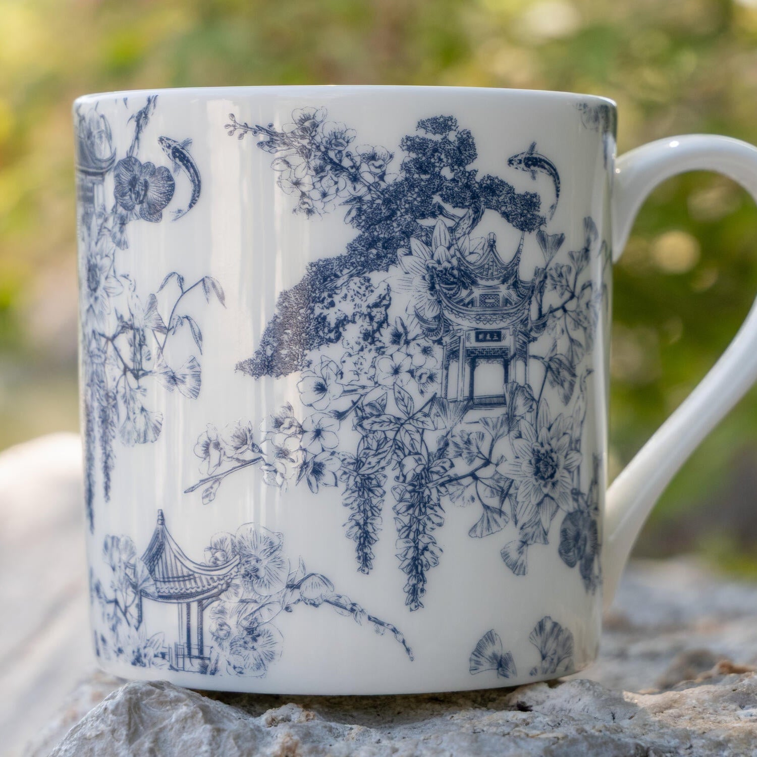 A white ceramic mug with a blue Asian gardens toile print, featuring pastel scenes and floral elements.