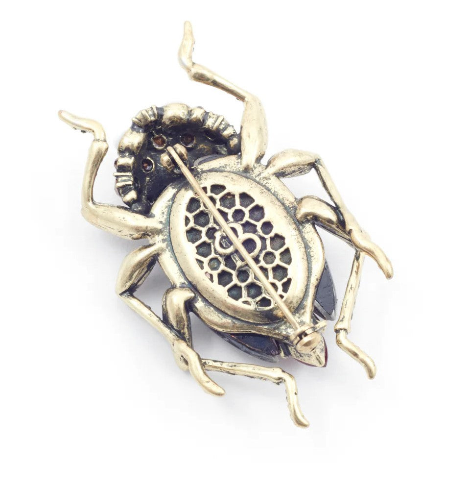 Add a finishing touch to your outfit with this bejeweled bug brooch.  This showstopper features bespoke resin stones and hand-set crystals meticulously crafted to form the body of this beautiful beetle. Materials: Brass with antique gold plating. Acrylic, resin, crystal Dimensions: 2.3" x 1.4". Pin fastening.