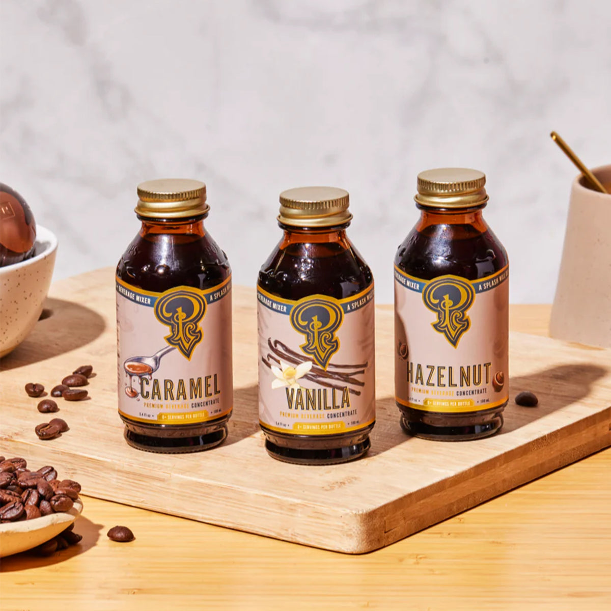 Three small amber bottles of coffee-flavored drink mix labeled Caramel, Vanilla, and Hazelnut, arranged on a wooden board with coffee beans and a marble background.