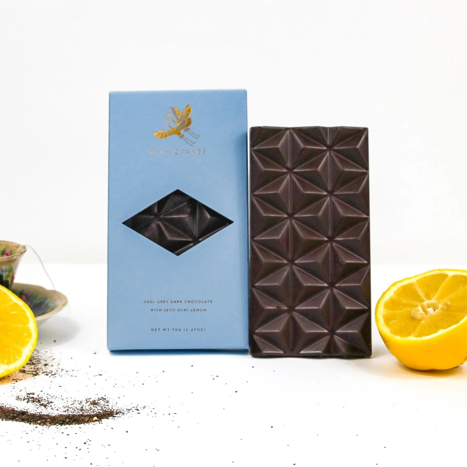 A light blue rectangular box of Deux Cranes Earl Grey dark chocolate, featuring a gold leaf logo and a diamond-shaped cutout window showing dark chocolate inside, set against a plain white background.