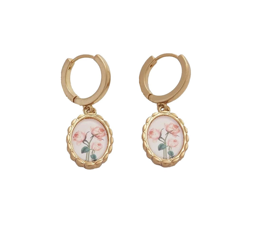 Experience the allure of Victorian elegance with these Framed Roses Huggies. These delicate pendants, featuring Dr Robert John Thornton's painted rose illustrations, hang from lobe-hugging hoops set in a timeless gold-tone oval hand-carved frame.
