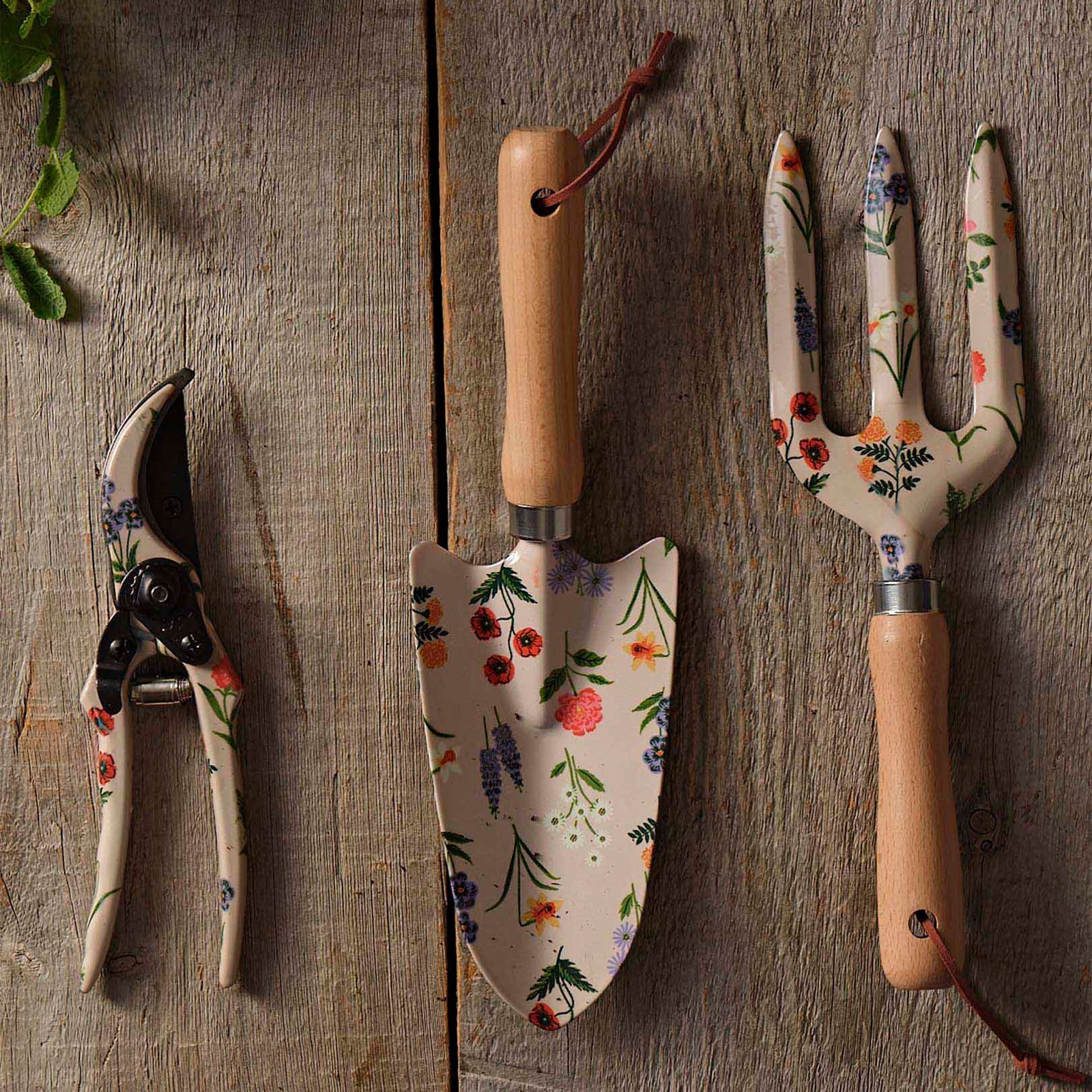 Three gardening hand tools with floral enamel handles and blades, including a garden fork, pruning shears, and trowel, shown on a plain white background.