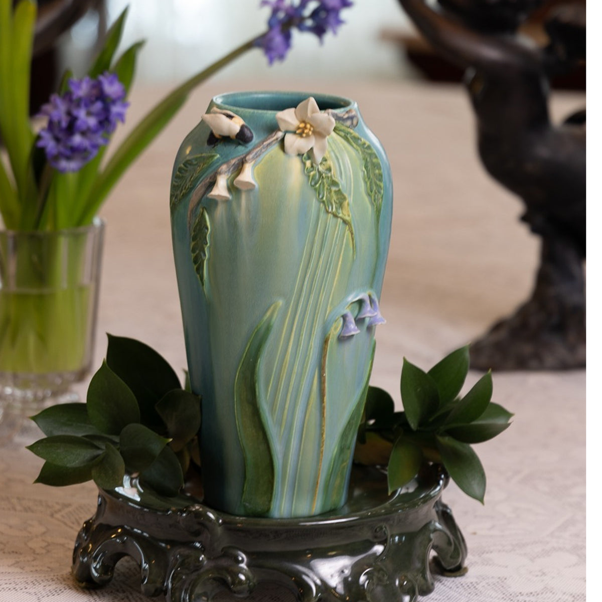 Decorative ceramic vase with floral design on a stand, surrounded by greenery and flowers.