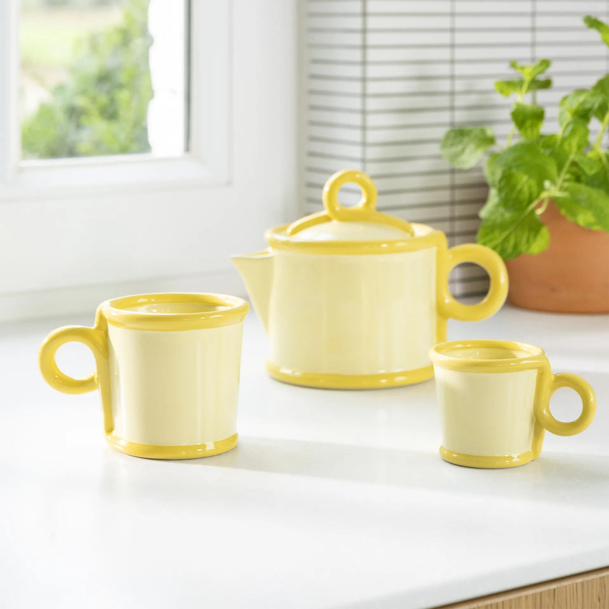 Yellow ceramic teapot and two mugs on a white surface with a window and plant in the background