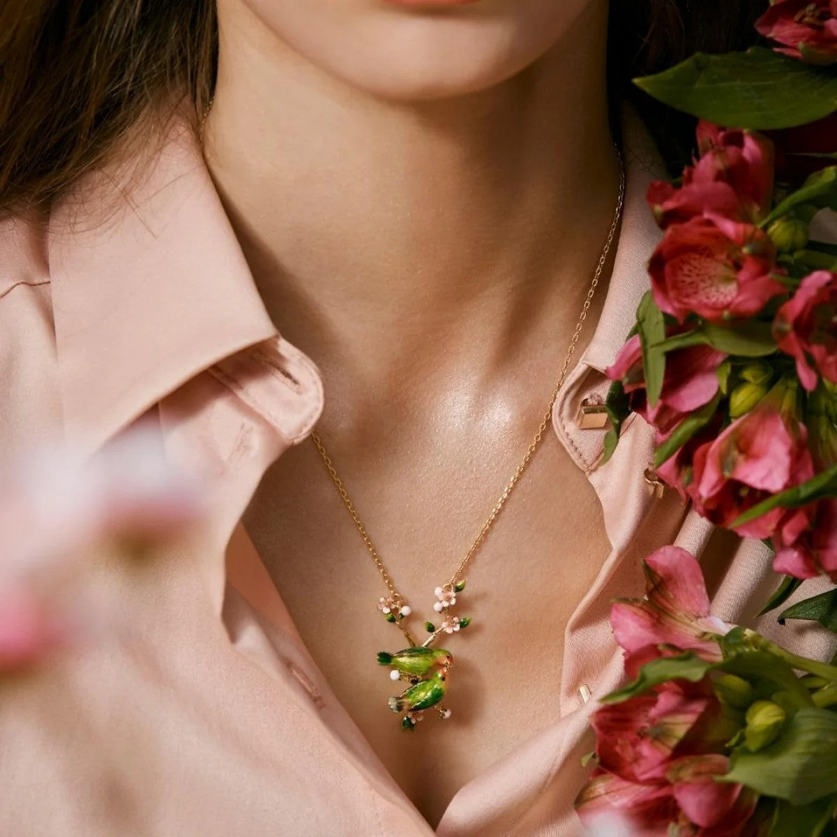 Person wearing a gold necklace with a green lovebird pendant, holding pink flowers.