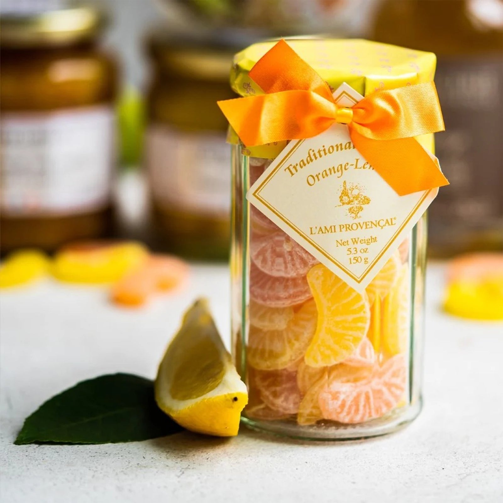 A clear glass jar filled with orange and yellow dried citrus candies, tied with a bright orange ribbon and a diamond-shaped label that reads 'Traditional Candies Orange-Lemon L'ami Provençal'.