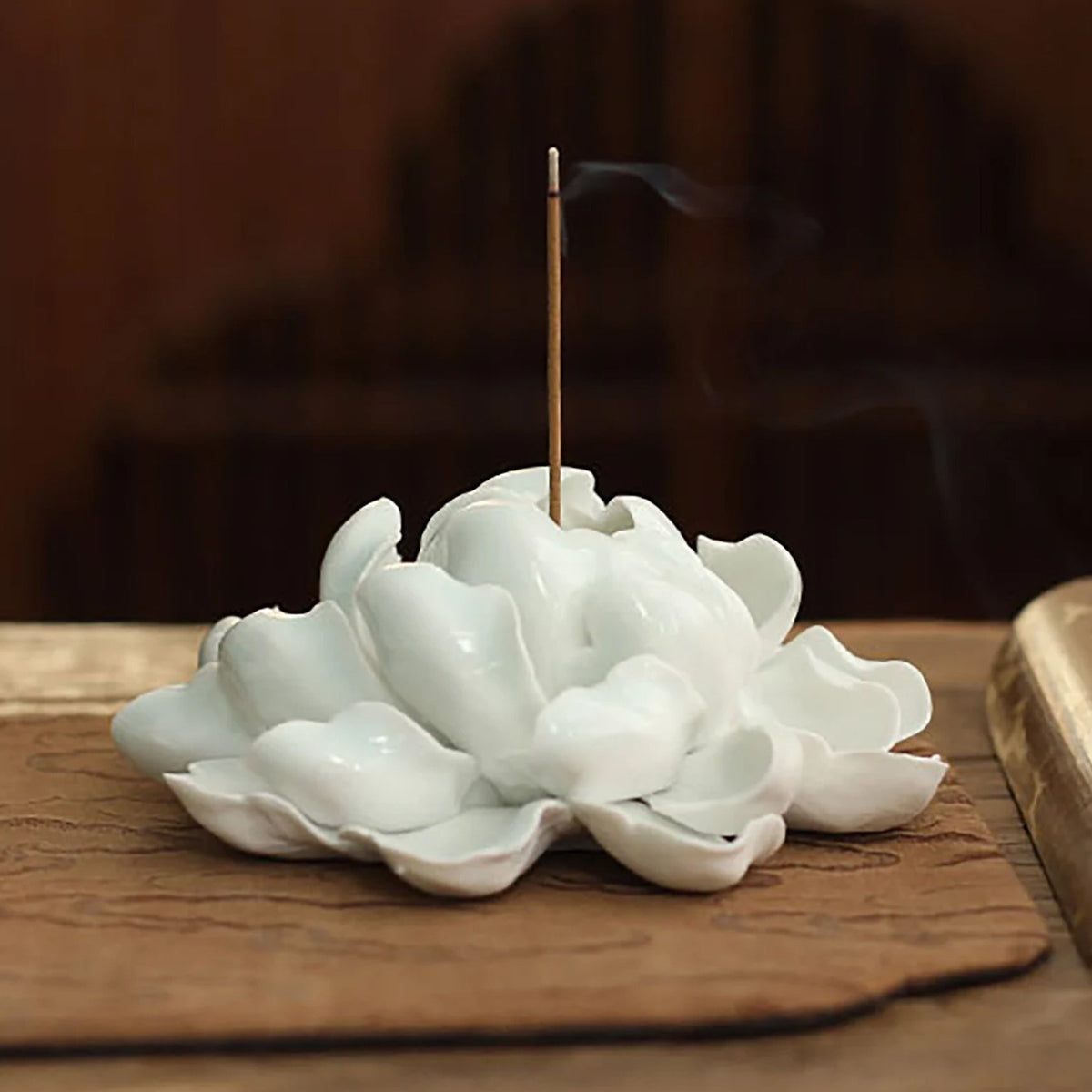 White ceramic incense burner shaped like a peony flower with a smoking incense stick on a wooden surface.