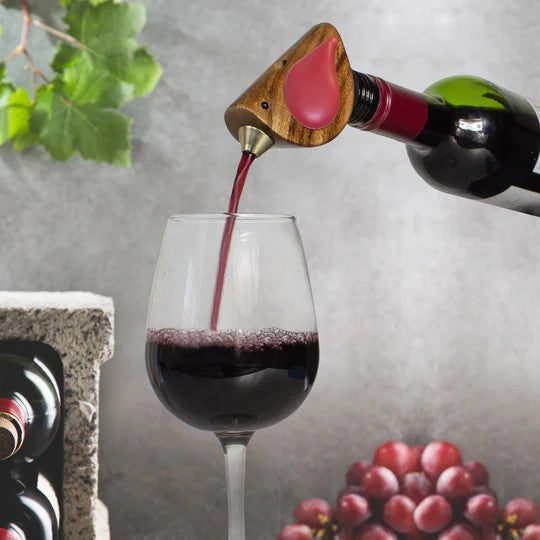 Red wine being poured into a glass with a decorative bottle stopper, grapes, and a stone surface in the background.