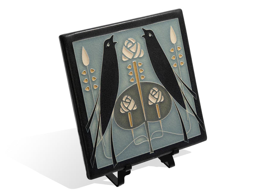 A handcrafted ceramic tile featuring two black birds and a rose design in beige and darker shades on a light blue background.