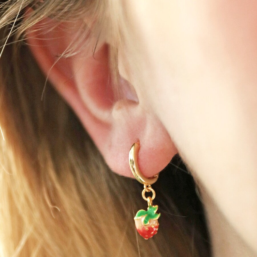 A delicate pair of gold huggie hoop earrings featuring dangling red strawberry charms, made from enamel. Designed with a simple click-in-place fastening, these adorable earrings are ideal for wearing every day to bring color and fun to an outfit. Materials: 14ct gold plated brass, enamel. 1" x 0.25".