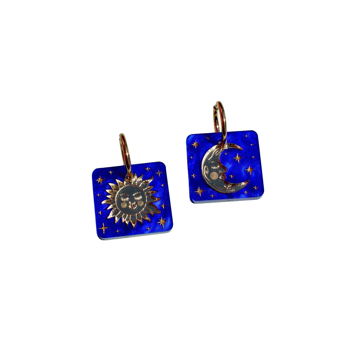 A pair of square cobalt blue glass earrings with gold-toned metal hoops featuring a sun face on one charm and a crescent moon with stars on the other, set against a plain white background.