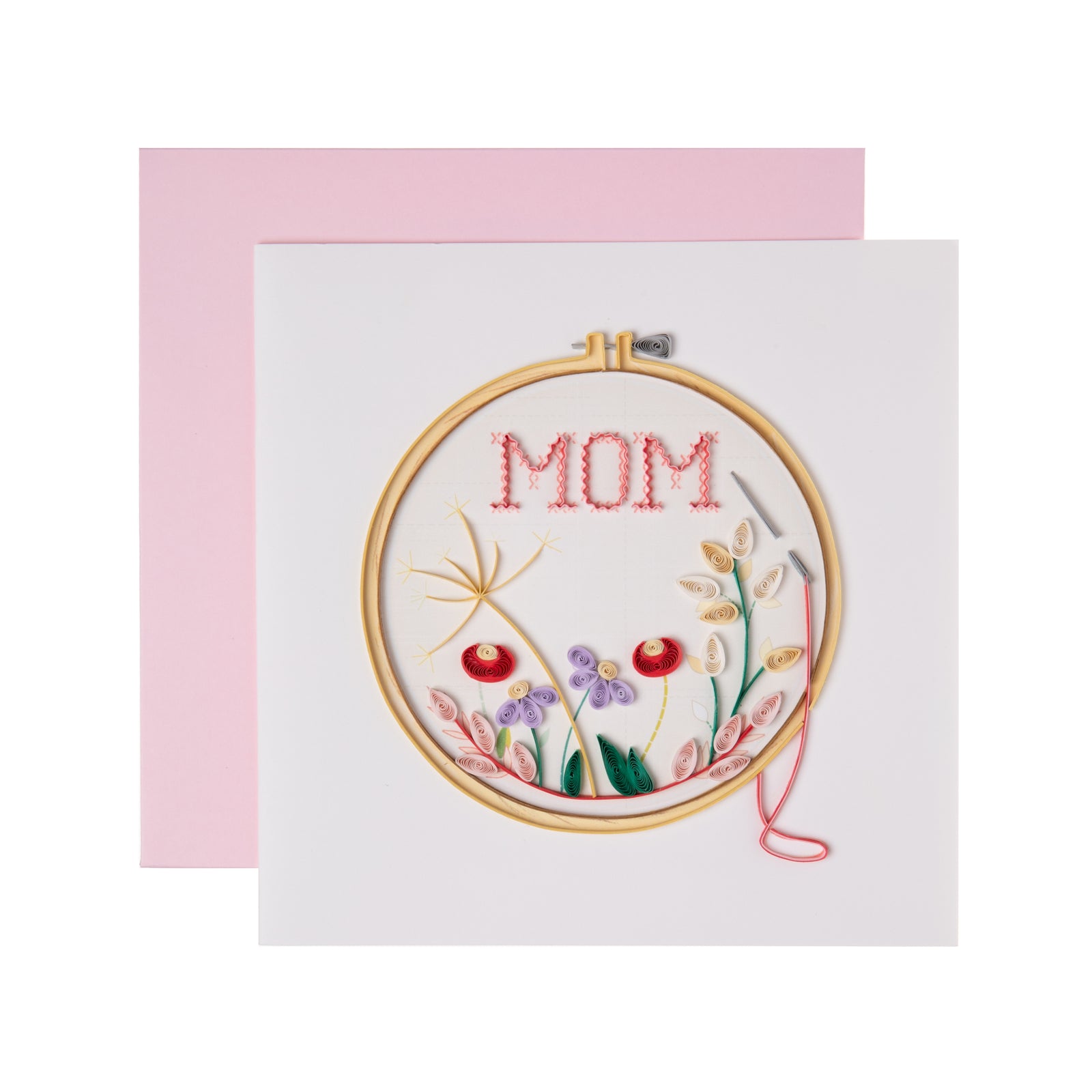 A greeting card with a quilled cross-stitch design featuring the word 'MOM' and floral patterns in red, purple, pink, and yellow on a hoop, enclosed in a pink envelope.