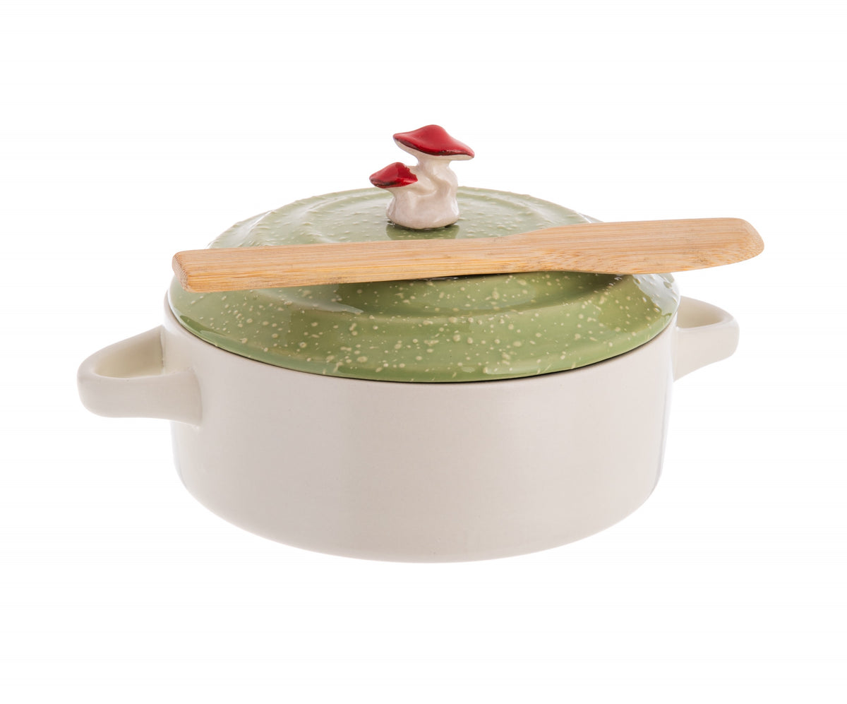 A white ceramic brie baking dish with a green lid adorned with a whimsical red field mushroom, accompanied by a bamboo spreader.