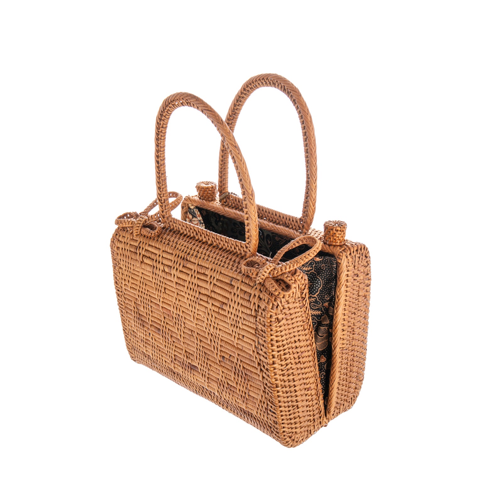 A woven rattan handbag with a brown color, featuring two bow clasps on top for closure, and a Batik print lining on the inside. It has a leather strap and hardware for handling.