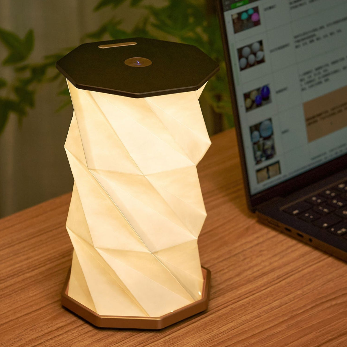 A modern geometric LED table lamp with a faceted, folded paper-like design, glowing with a warm cream light. It has a matte black top and base with a small circular metallic button on top.