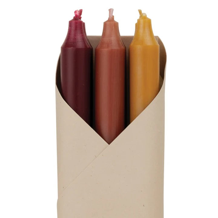 Set of six candles in red, brown, and yellow in a cardboard holder with a label on a white background.