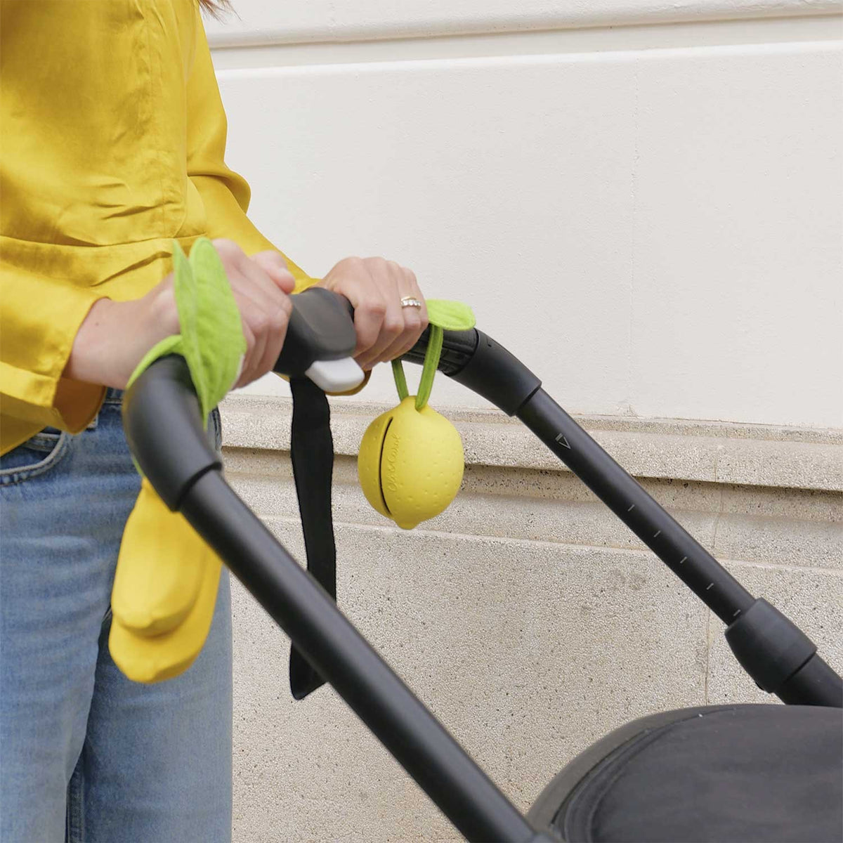 Person pushing a stroller with a yellow lemon-shaped pacifier case attached, wearing a yellow jacket and blue jeans.