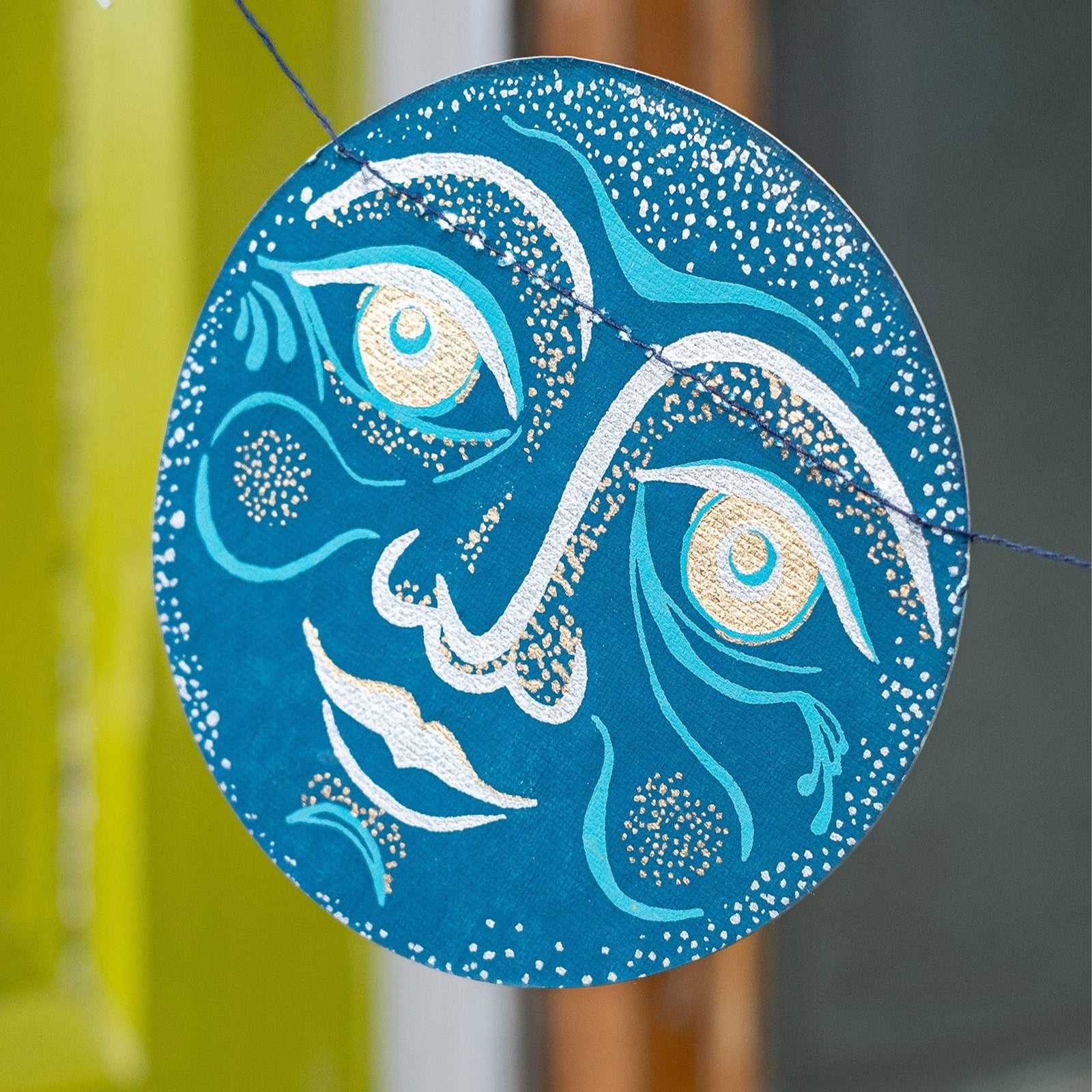 A celestial-themed garland banner featuring painted moon and star shapes in teal, blue, beige, and silver, strung along a thin white cord against a plain white background.