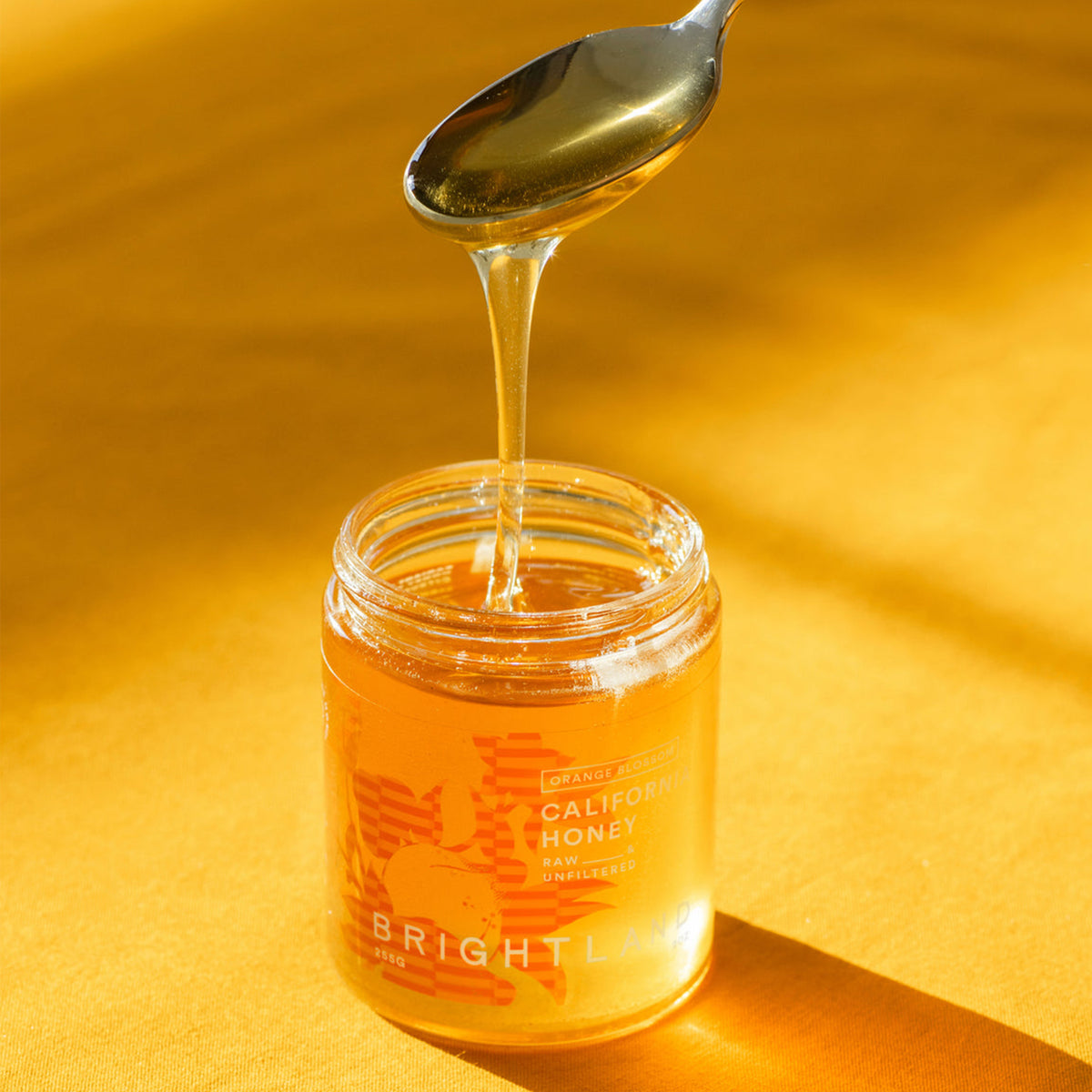 A jar of Bright Lake California Honey with golden honey dripping from a spoon against a warm yellow background.
