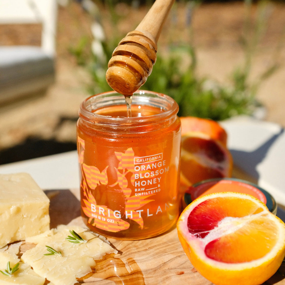 A jar of Brightla orange blossom honey sits on a wooden board with a honey dipper above it, surrounded by peach halves, a slice of pale cheese, and a halved orange in a bright outdoor setting.