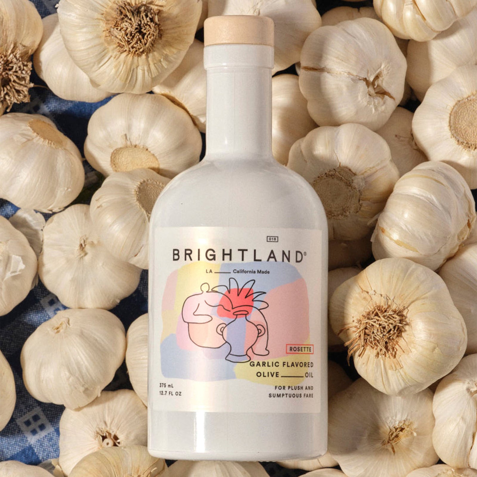 A white glass bottle of Brightland Garlic Flavored Olive Oil with a light wood cap and a colorful label featuring a stylized face illustration.