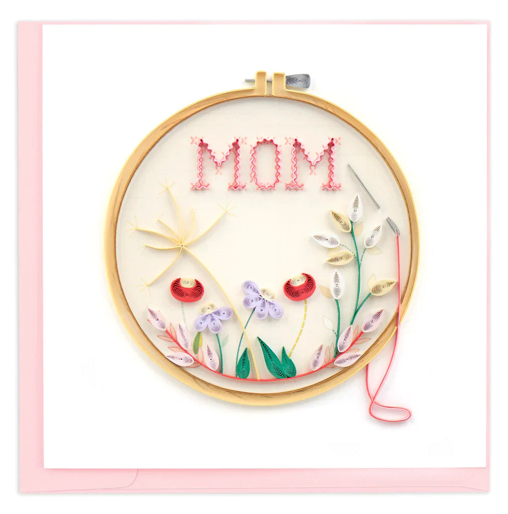 A greeting card with a quilled cross-stitch design featuring the word 'MOM' and floral patterns in red, purple, pink, and yellow on a hoop, enclosed in a pink envelope.
